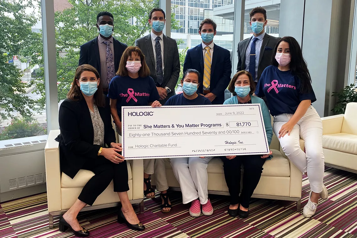 Gruppefoto af maskerede personer, der holder en stor symbolsk check til støtte for programmerne She Matters og You Matter.